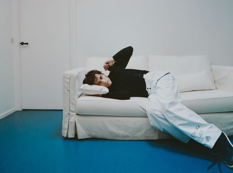 Man lying down on a white fabric sofa wearing a black long-sleeve shirt and white pants on a blue floor.