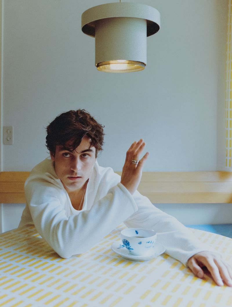 Portrait of a young man with dark hair in white top sitting at a table with a cup and saucer in an indoor setting under a light fixture.