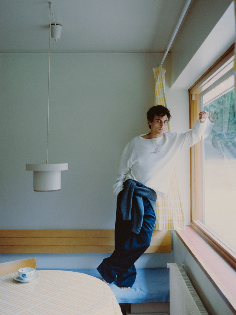Julien de Saint Jean leaning against a window frame in a white sweatshirt and dark trousers in a minimalist domestic room.