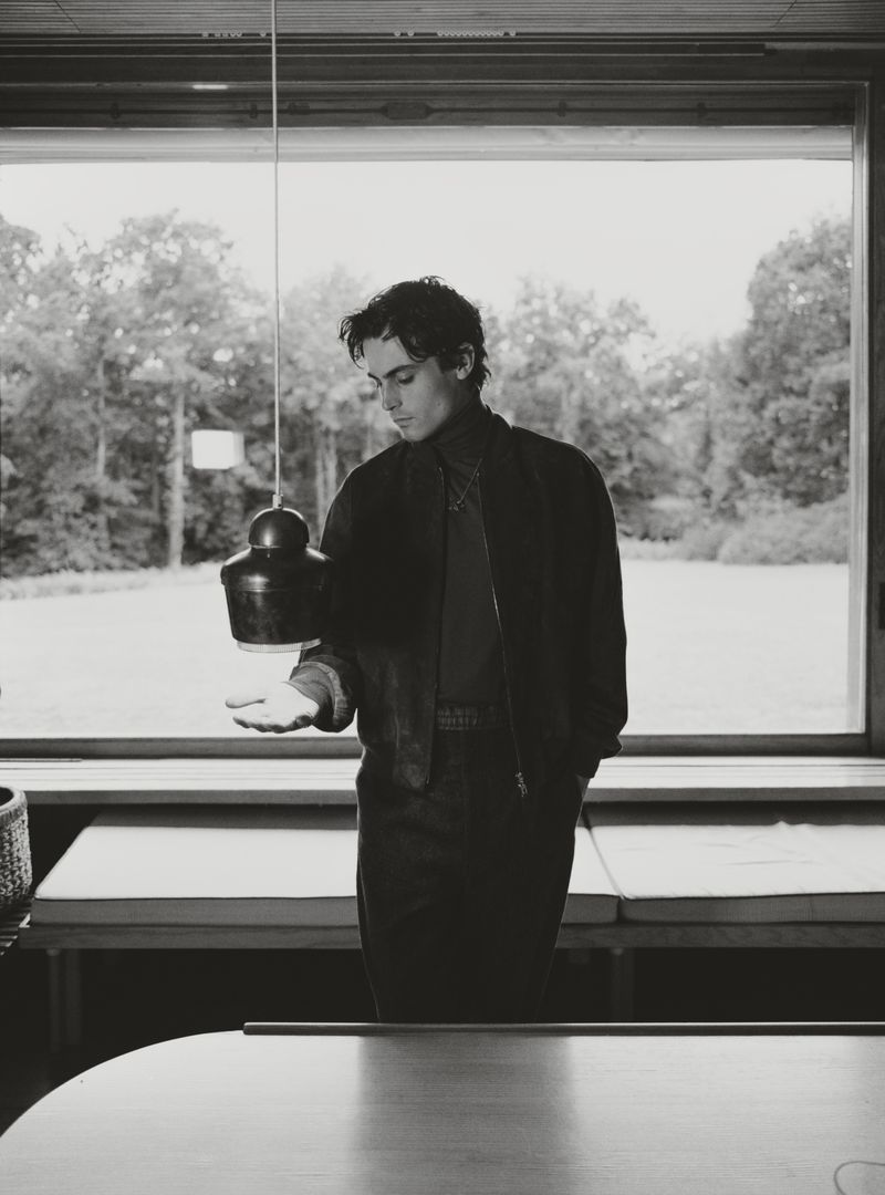 Black and white portrait of a young adult man in an interior setting, posing in front of a large window with trees visible outside.