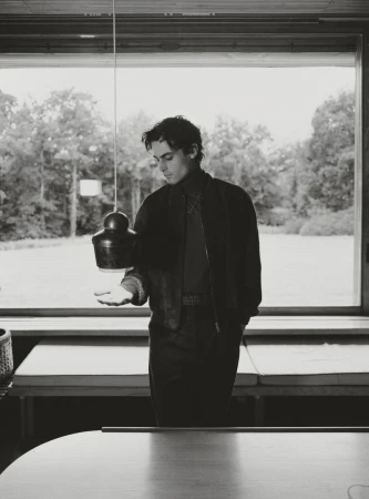 Black and white portrait of a young adult man in an interior setting, posing in front of a large window with trees visible outside.