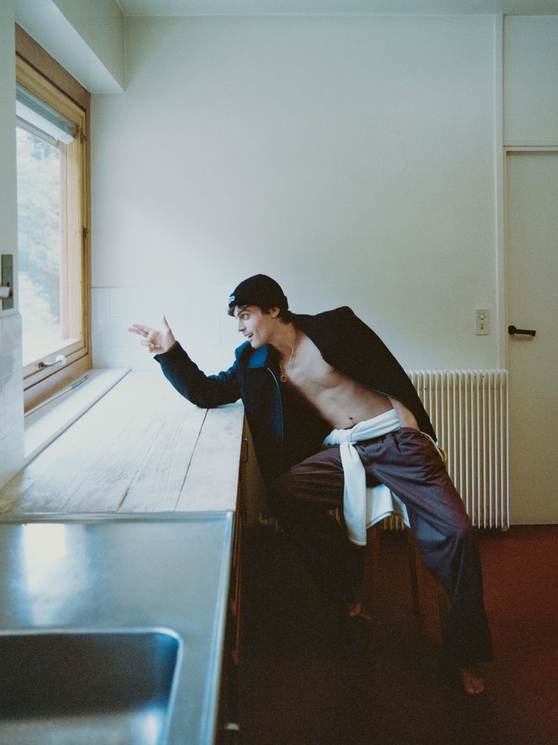 Bare-chested adult male posing near window in kitchen, wearing a black jacket and brown pants, looking to the side with finger pointing.