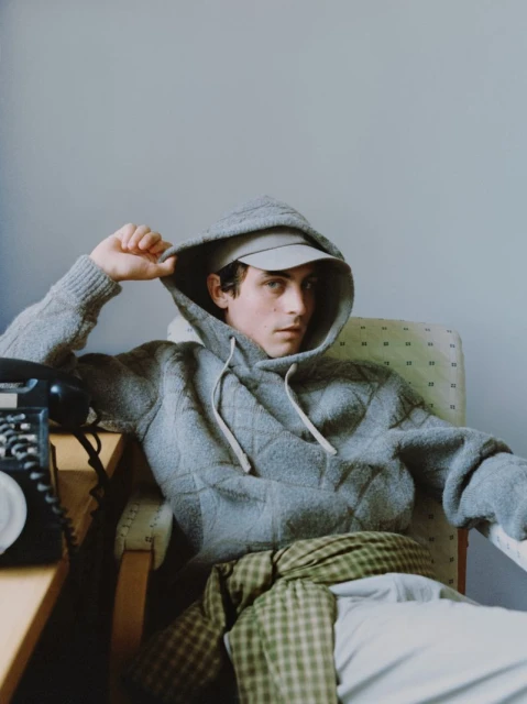 Man in a grey hoodie and cap sitting in a chair indoors, with a rotary phone on a desk next to him, captured in soft, natural light.
