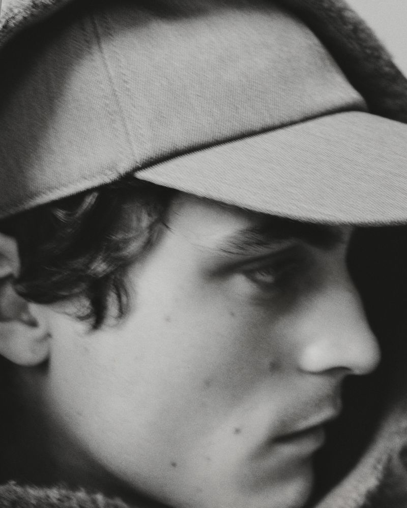 Black and white close-up portrait of a man wearing a baseball cap and hooded garment looking downward.
