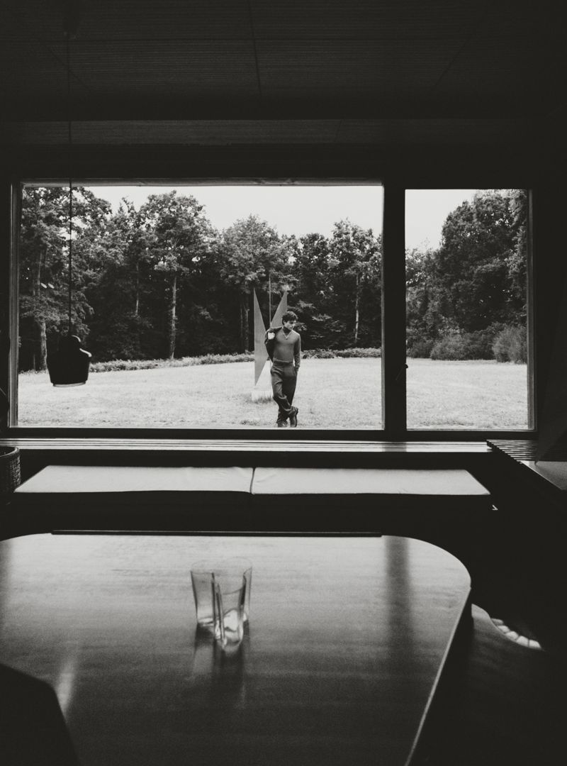 Monochrome interior view shows a person standing outside a window, alongside a modern sculpture, with glassware in the foreground.