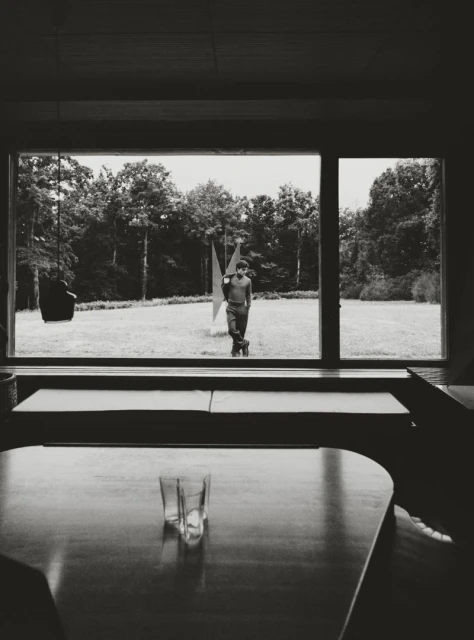 Monochrome interior view shows a person standing outside a window, alongside a modern sculpture, with glassware in the foreground.
