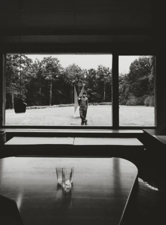 Monochrome interior view shows a person standing outside a window, alongside a modern sculpture, with glassware in the foreground.