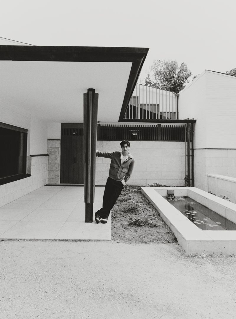 Monochrome shot of a man leaning against a pillar outside a modern building with minimal landscaping. He wears a jacket and dark pants.