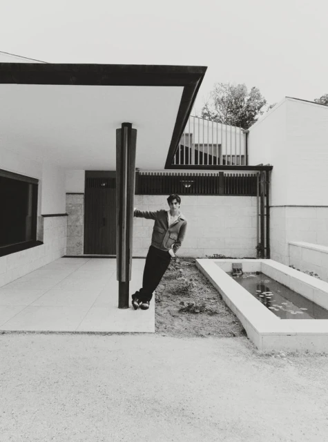 Monochrome shot of a man leaning against a pillar outside a modern building with minimal landscaping. He wears a jacket and dark pants.