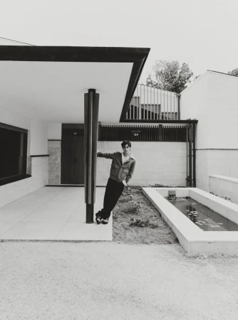 Monochrome shot of a man leaning against a pillar outside a modern building with minimal landscaping. He wears a jacket and dark pants.
