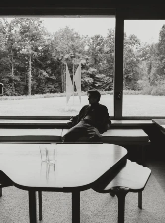 Black and white portrait of Julien de Saint Jean seated on a bench by a large panoramic window overlooking trees and a sculpture.
