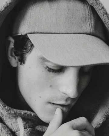 Monochrome close-up portrait of Julien de Saint Jean wearing a baseball cap and a textured hoodie looking downward.