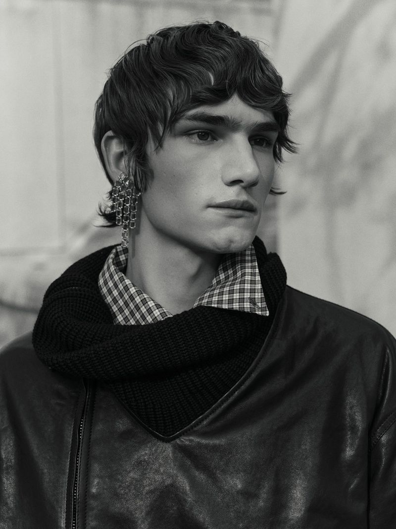 Monochrome portrait of a young adult male with dark hair wearing an earring, collared plaid shirt, knit scarf, and leather jacket.