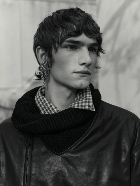 Monochrome portrait of a young adult male with dark hair wearing an earring, collared plaid shirt, knit scarf, and leather jacket.