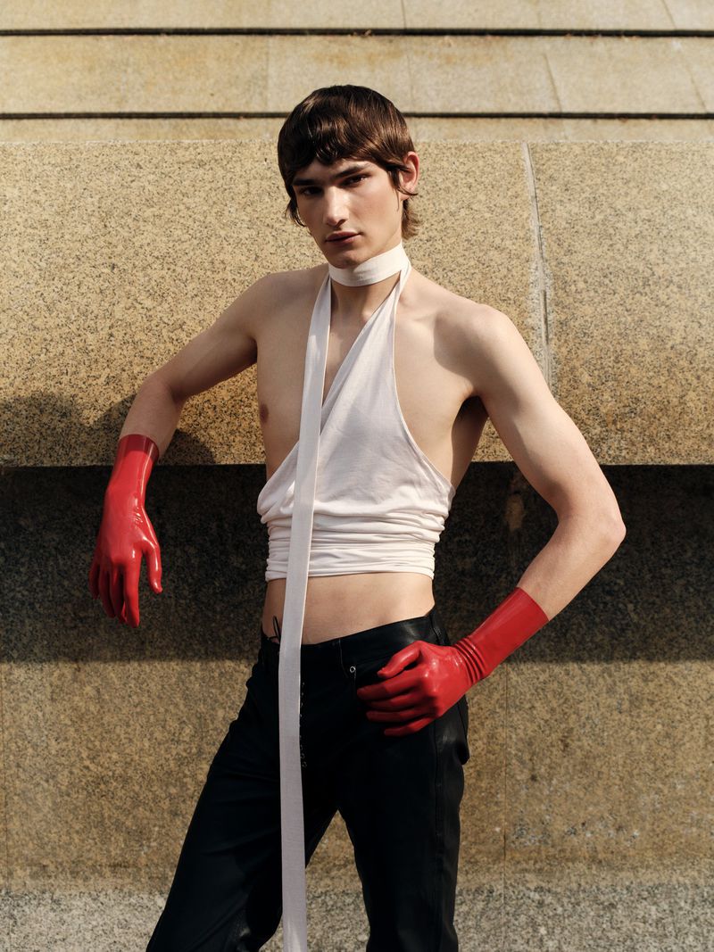 A slender young adult stands with hands on hips in front of a stone wall, posing in a minimalist white top and striking red gloves.