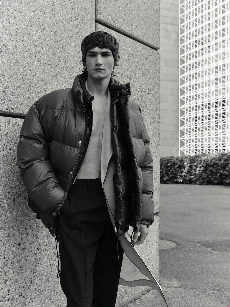 Male model poses in puffer jacket outdoors against concrete wall with architectural structure, wearing black trousers and open shirt in black and white.
