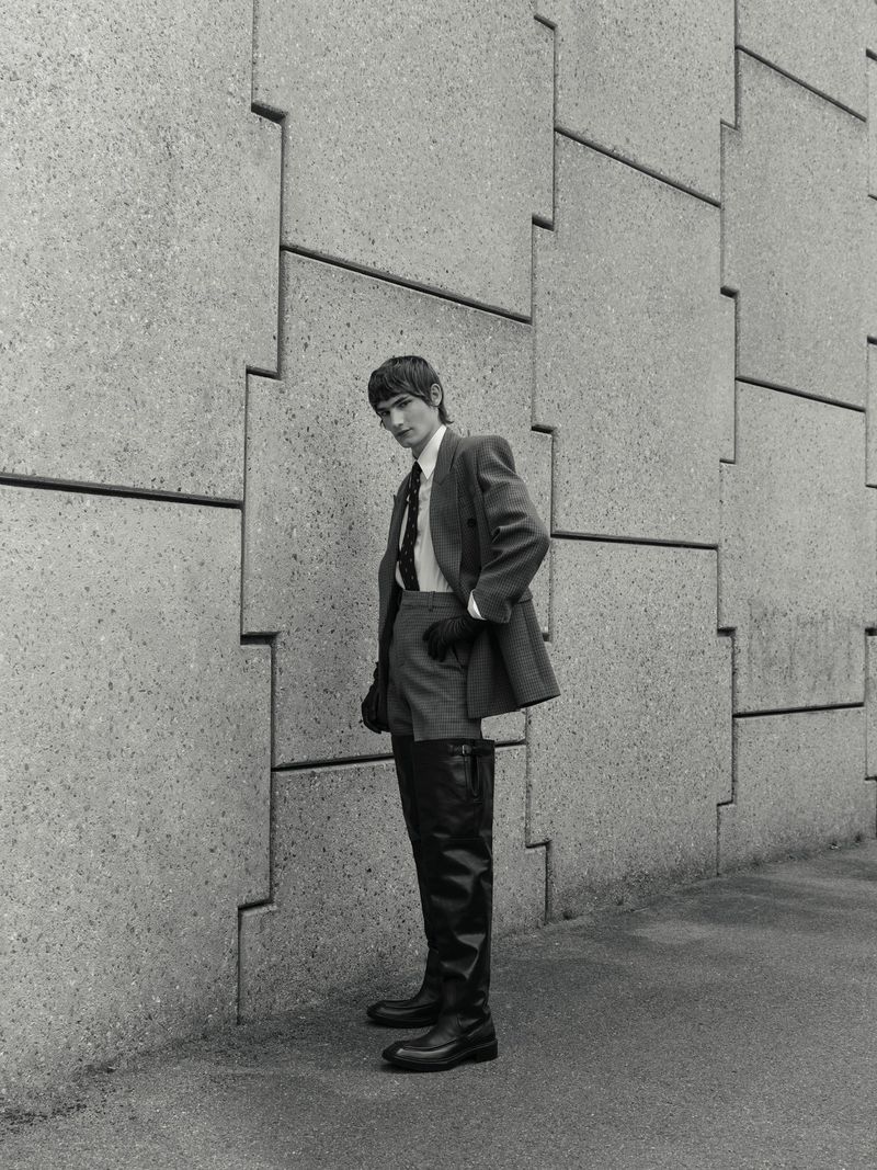 Model in grey suit jacket and tall dark boots leaning against a textured concrete wall outdoors.