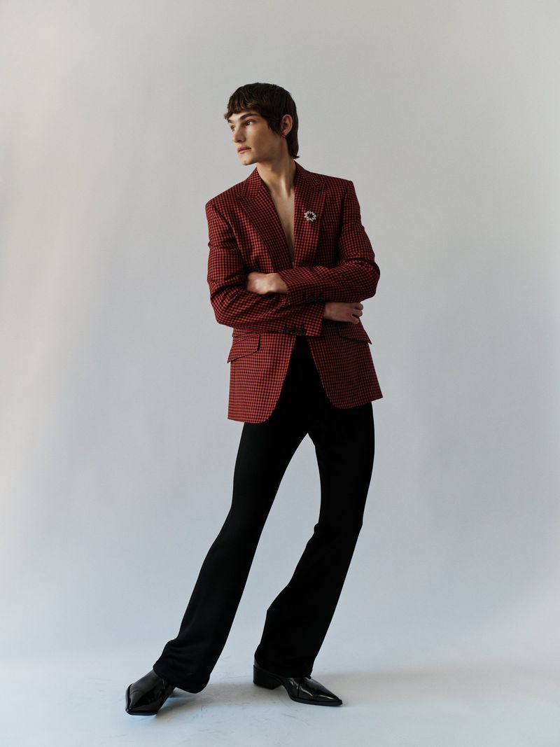 A slender male fashion model poses in a patterned blazer and black pants against a neutral background, looking to the side.