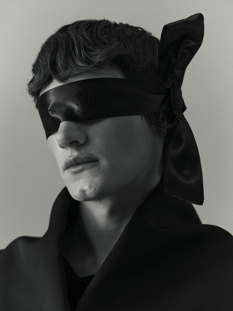 Monochrome portrait of a person with a black ribbon blindfold, wearing a dark coat, captured in a studio setting with soft lighting.