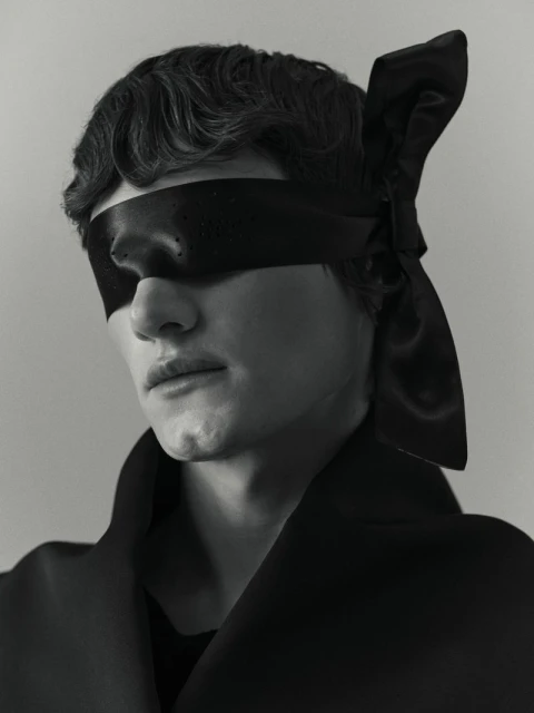 Monochrome portrait of a person with a black ribbon blindfold, wearing a dark coat, captured in a studio setting with soft lighting.