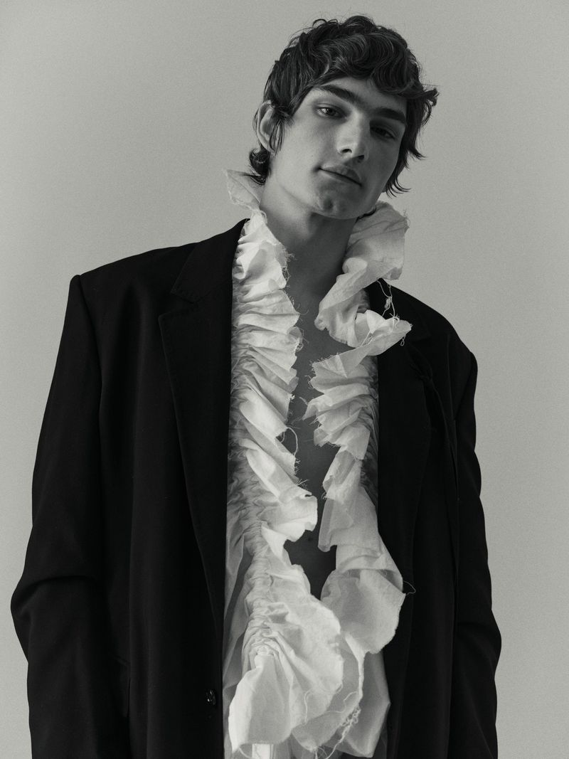 Monochrome portrait of a young adult male wearing a dark blazer and a ruffled white shirt in a studio setting.