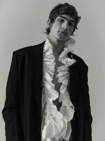 Monochrome portrait of a young adult male wearing a dark blazer and a ruffled white shirt in a studio setting.