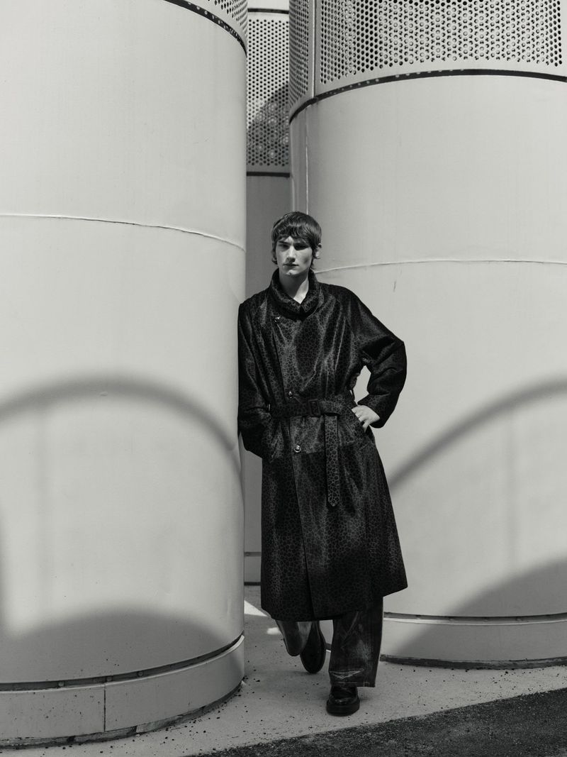 Monochrome portrait of a male model in a long patterned coat, leaning against a concrete structure. Fashion editorial style.