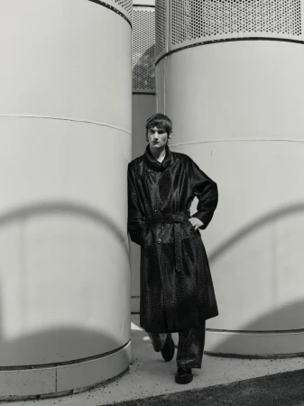 Monochrome portrait of a male model in a long patterned coat, leaning against a concrete structure. Fashion editorial style.