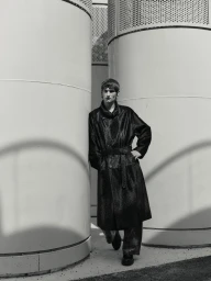 Model Silas Lutz Fabian wearing a dark belted long coat standing between large curved metal architectural columns in black and white.