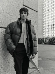 Silas Lutz Fabian wearing an oversized black puffer jacket and trousers outdoors against an architectural wall for Behind the Blinds.