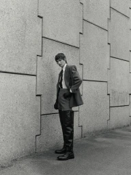Model in grey suit jacket and tall dark boots leaning against a textured concrete wall outdoors.
