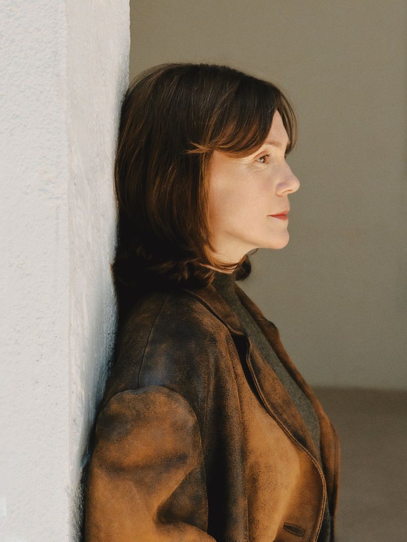 Profile portrait of a woman leaning against a textured white wall wearing a brown distressed leather jacket