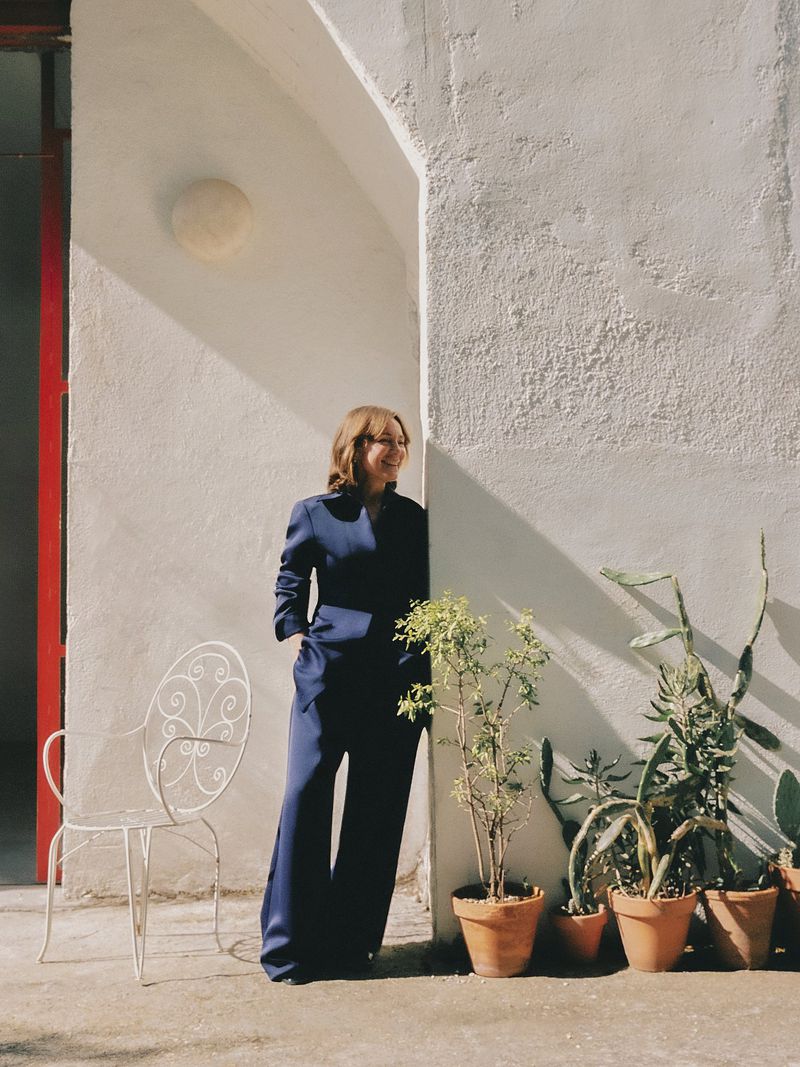 A woman in a navy suit stands next to potted plants in an outdoor setting with white walls and shadows, looking off to the side.