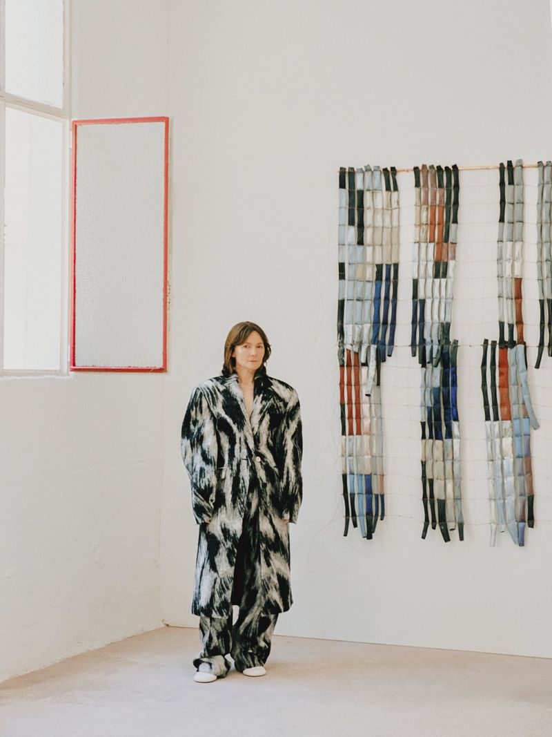 A woman in a black and white patterned coat and pants standing in a white corner near an art installation and a window.