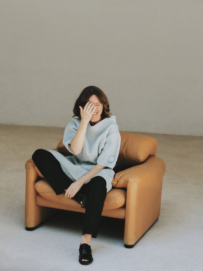 Adult female in oversized sweater and black pants sits in a tan leather chair with a hand to her face in a minimal indoor setting.