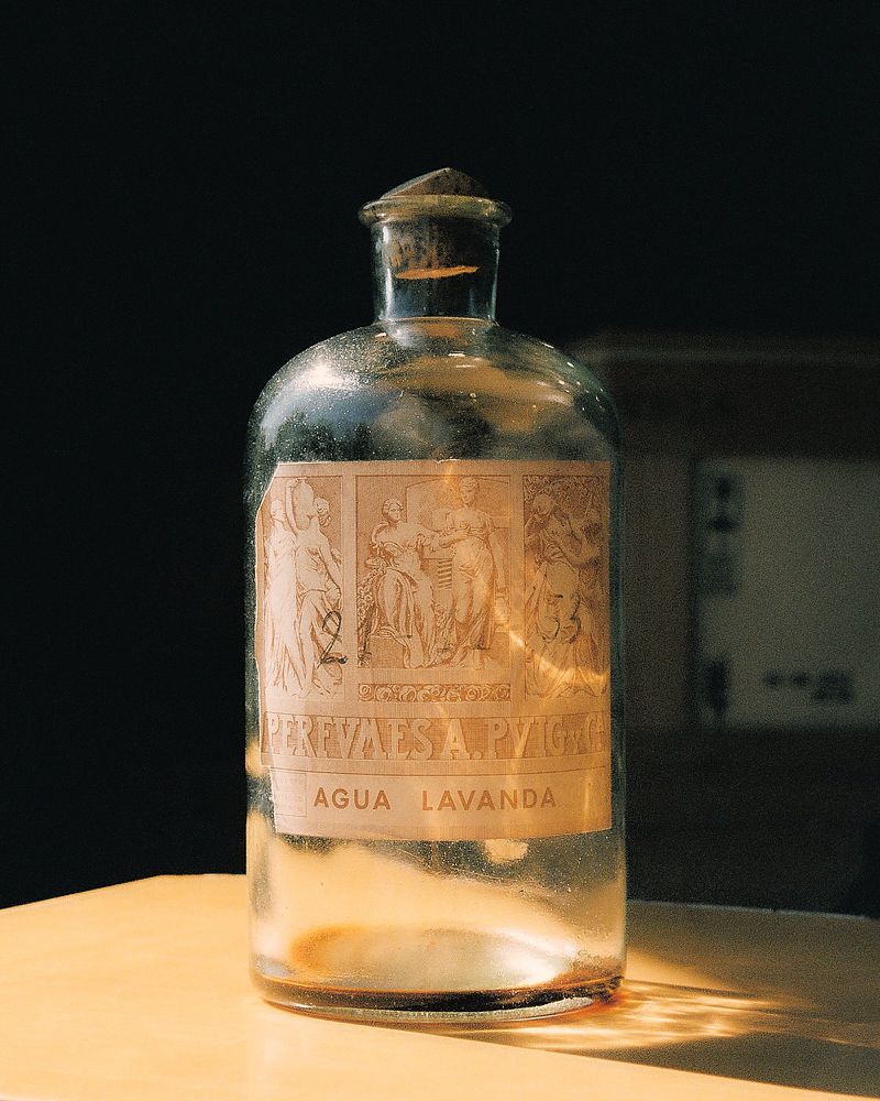 Antique clear glass bottle filled with liquid, featuring a decorative label with classical figures, casting shadows on a tabletop.
