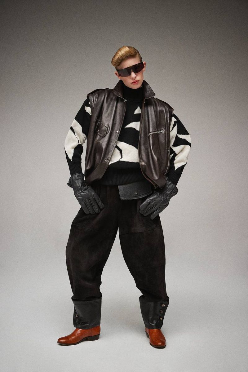 Male model in a black and white sweater vest and oversized black trousers poses in a studio with tan leather boots and gloves.