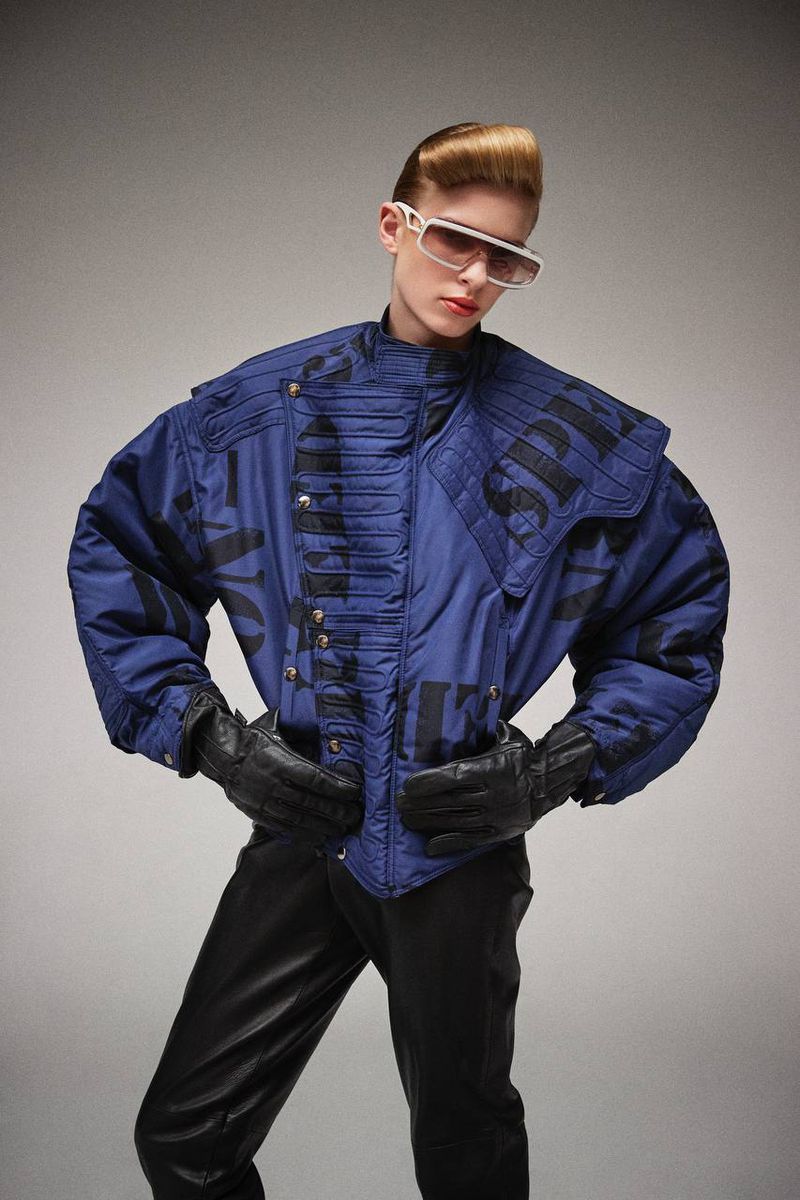 Person in blue jacket with black lettering, black leather pants, and white sunglasses poses in a photo studio. Fashion shot.