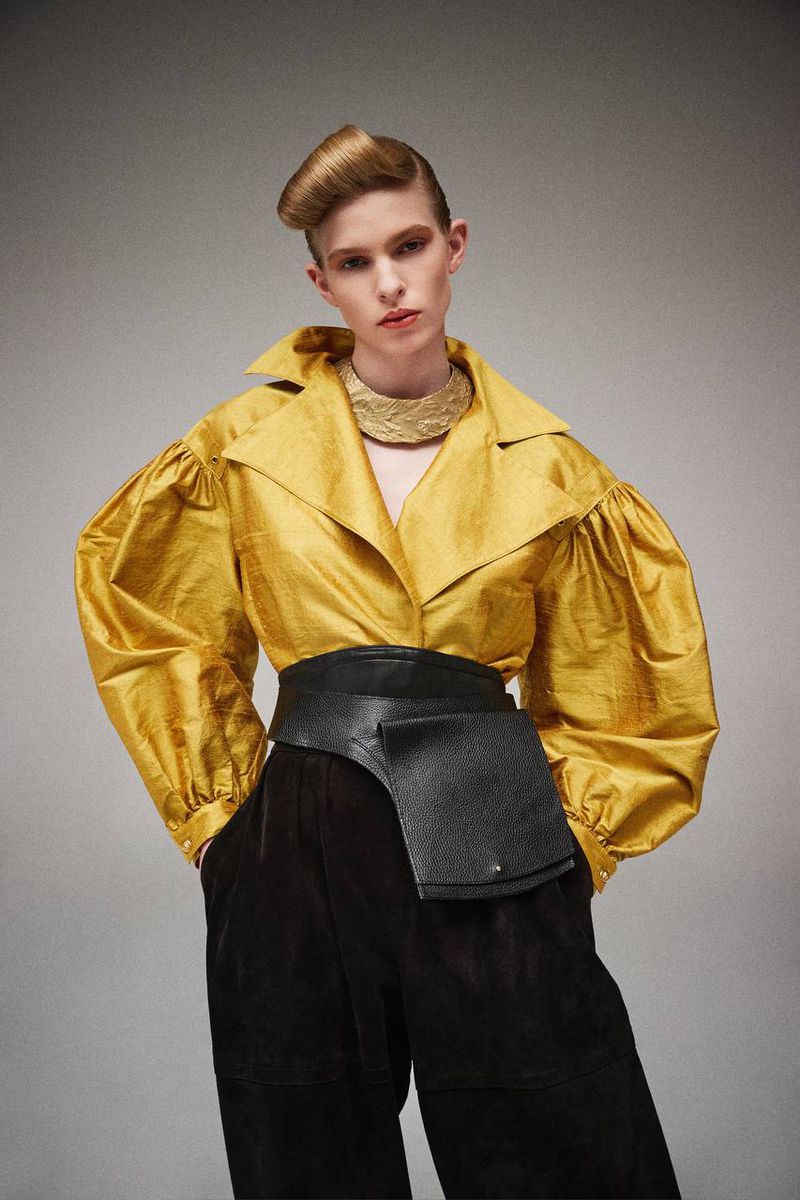 Woman in gold silk jacket with balloon sleeves and black wide belt stands in studio. Haute couture fashion portrait, studio shot.