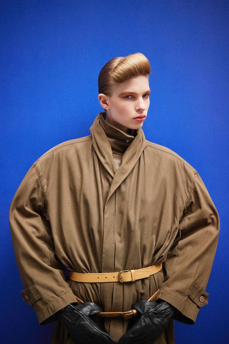 Fashionable person with pompadour hairstyle in tan trench coat with black gloves against a blue background in a studio setting.