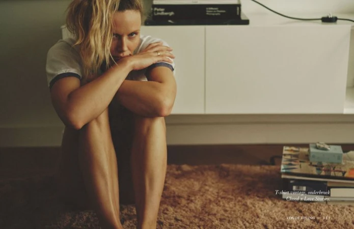 Indoor portrait of a fair-skinned woman with blonde hair sitting on the floor next to white furniture and books, looking at the camera.