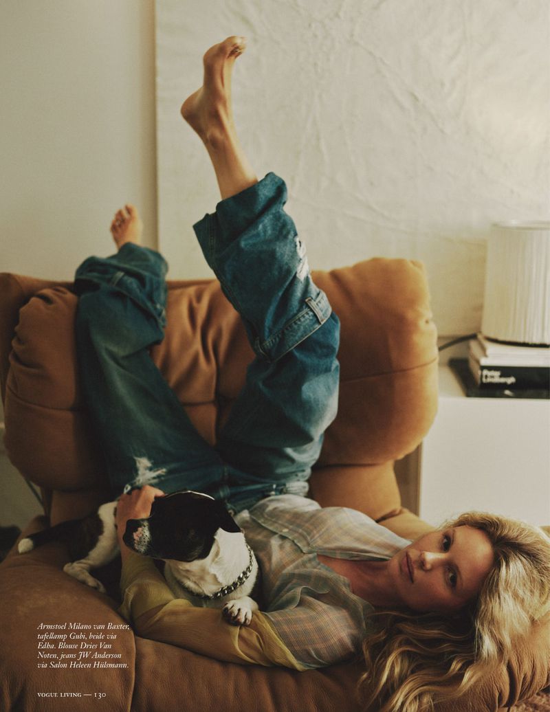 Adult woman reclines on a brown chair indoors with her dog, wearing jeans and a light shirt, relaxed pose and casual setting.