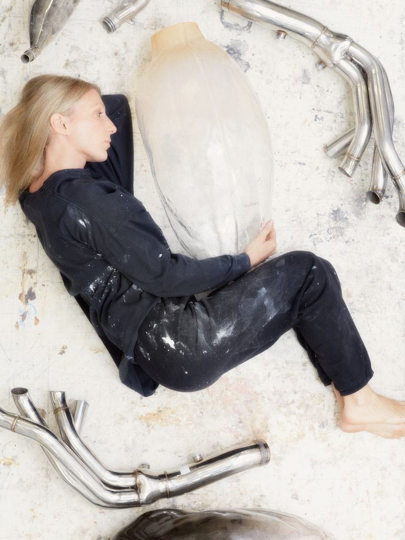 An adult woman lies on a light surface with a large wrapped object and chrome pipes scattered around her in an indoor environment.