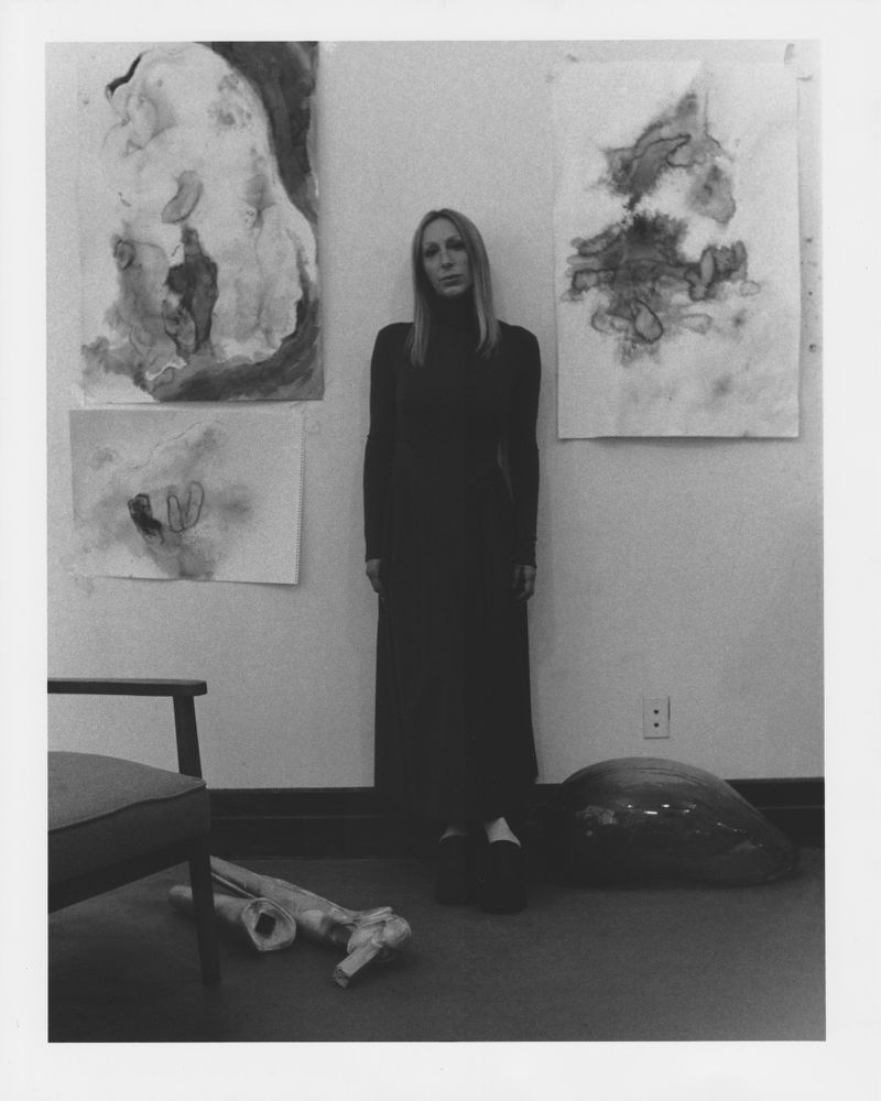 Black and white portrait of Ivana Bašić standing in a black dress between sketches on a wall for Vogue Adria.