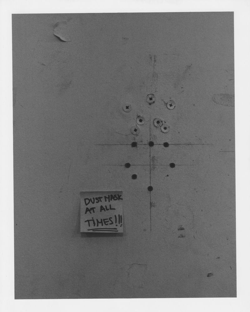 Image of a wall with a note attached reading 'DUST MASK AT ALL TIMES!!!', surrounded by screws and markings, showcasing a utilitarian aesthetic.