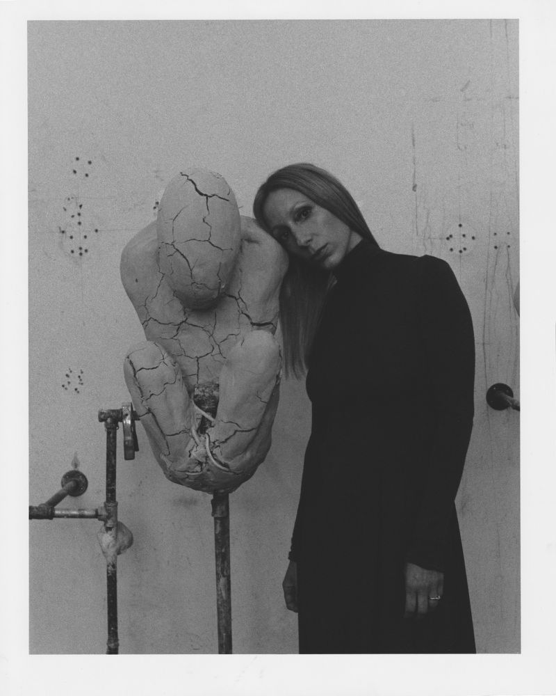 Monochrome portrait of a blonde woman resting her head on a cracked sculpture against a textured wall with metal pipes, creating a contemplative scene.