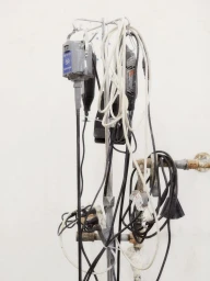 Tangled power cords and rotary tools hanging from a metal stand against a white wall with industrial pipes.