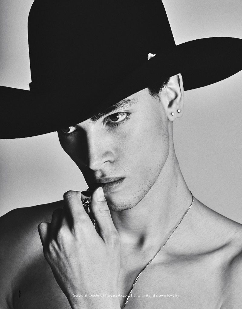 Black and white portrait of a young adult man wearing a black hat and silver chain, posing in a photo studio with soft, diffused lighting.