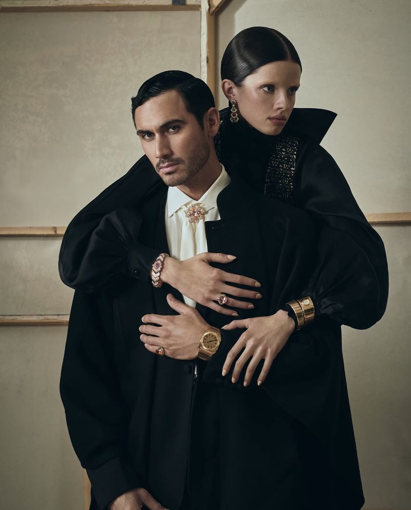 Elegant fashion shot of a couple, the woman's hands resting on the man's chest. Both wear black jackets and stylish gold jewelry.