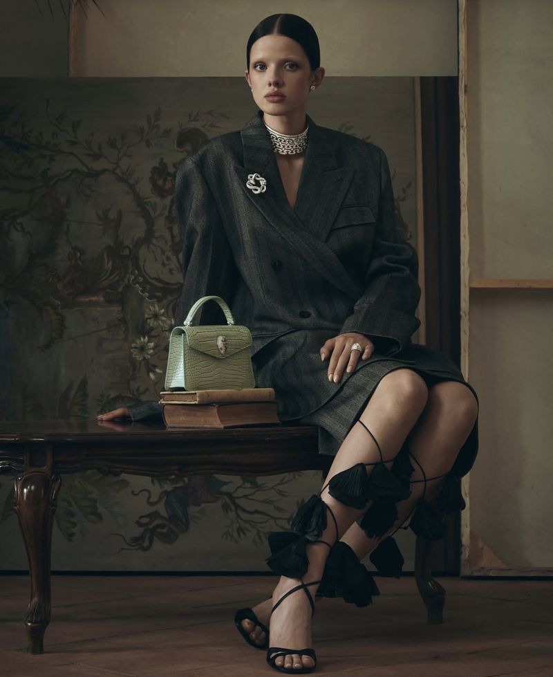 Fashion shot of a woman in a suit with a handbag sitting on a table in front of a painted canvas backdrop, wearing jewelry.